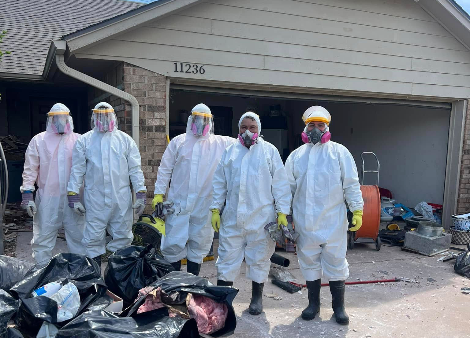 Five individuals in protective gear stand outside a house, surrounded by garbage bags in a cluttered area.