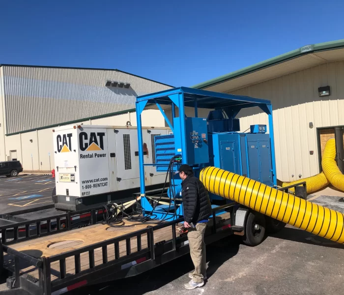 A worker inspects a blue power generator trailer with a yellow exhaust hose outside a large industrial building under a clear sky.