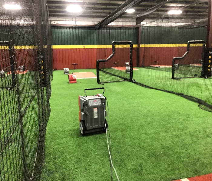 Indoor batting cage facility featuring turf flooring, training equipment, and netting for practice. Batting area with pitching machines visible.