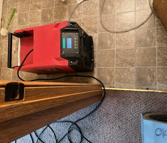 An overhead view of a red device with a digital display and multiple cables, resting on a tiled floor near wooden trim and carpet.