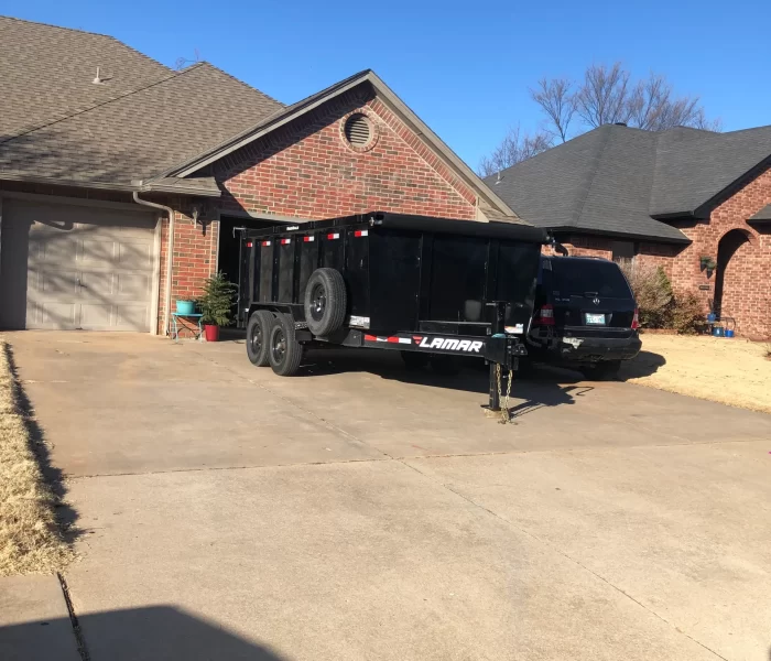 A black dump trailer parked in a driveway beside a dark SUV, with a brick house and clear blue sky in the background.