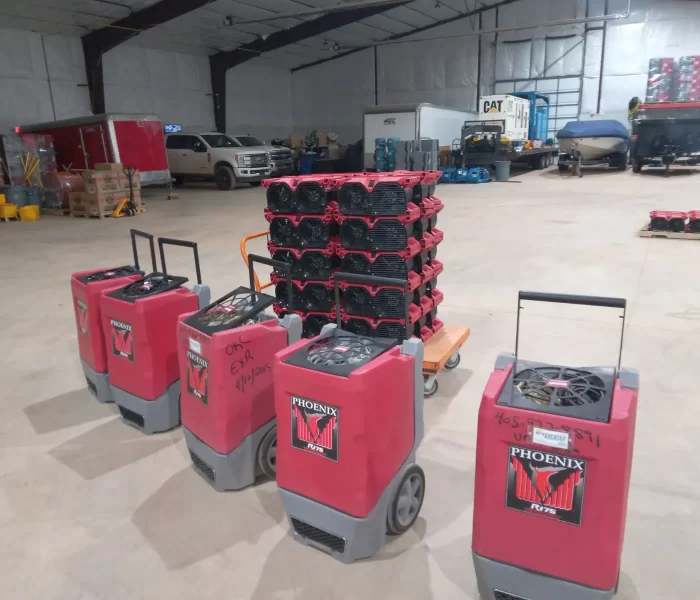 Four red Phoenix dehumidifiers are lined up in a warehouse, with stacked equipment and vehicles visible in the background.