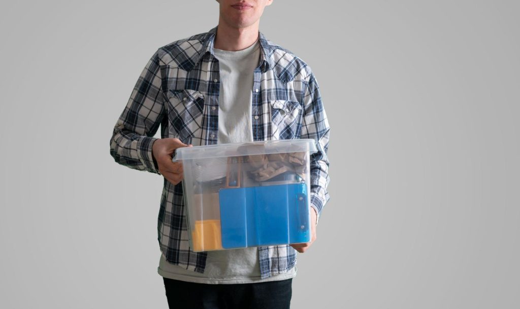 A young man is carrying a plastic storage box with items.