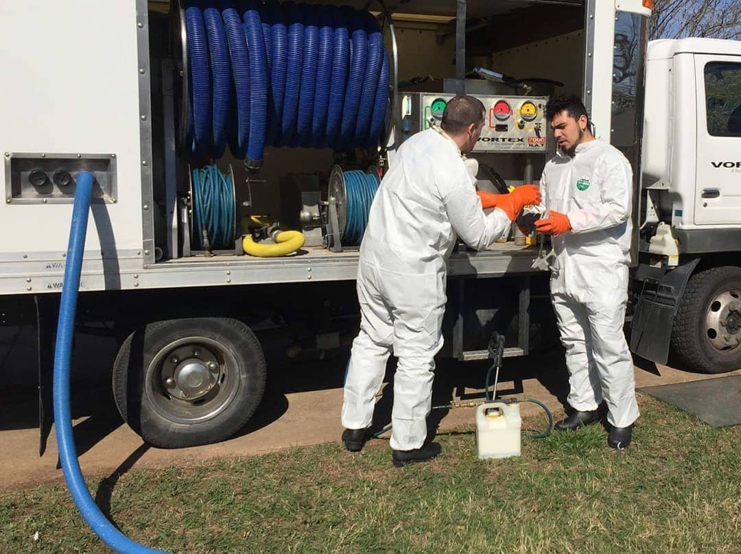 Two workers in white protective suits and orange gloves are interacting near machinery on a truck in a grassy area.